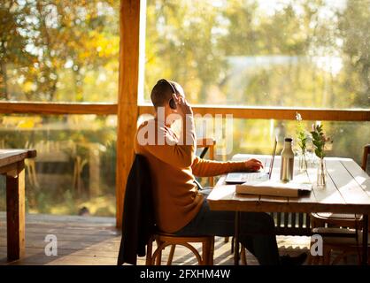 Uomo d'affari con le cuffie che lavorano al computer portatile nel soleggiato caffè autunnale Foto Stock