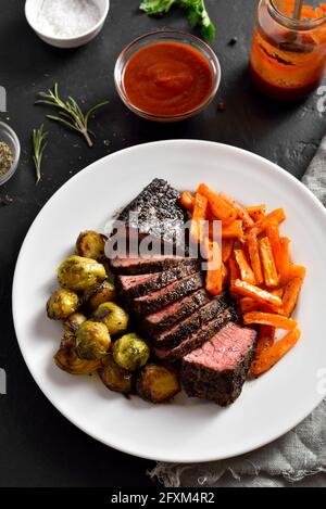 Bistecca di manzo alla griglia con germogli di bruxelles e patate dolci sul piatto su fondo di pietra nera. Vista ravvicinata Foto Stock