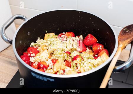 Fragole e fiori di sambuco marmellata cottura in pentola nera con cucchiaio di legno. La foraggazione primaverile conserva l'ispirazione della ricetta. Primo piano, vista laterale. Foto Stock