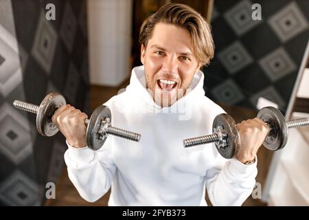 Ritratto di giovane attraente gioioso atletico caucasico che si allenano in palestra, appoggiandosi su manubri, mostrando muscoli del corpo, facendo allenamento dei bicipiti Foto Stock