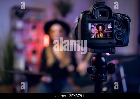 Sfocatura sfondo di musicista femminile che suona batteria elettronica e registrare video. Messa a fuoco della fotocamera digitale sul treppiede. Processo creativo in interni. Foto Stock