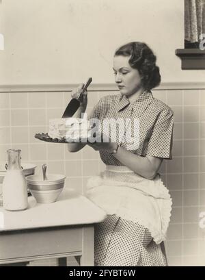 Una donna adulta, una casalinga o una cameriera, con una sciarpa verde  sulla testa e un grembiule rosso, si trova dietro l'asse da stiro. Lei ne  fa un po' Foto stock 