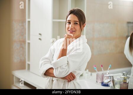 Elegante donna di charme in morbido accappatoio che tiene la mano sotto mento in bagno Foto Stock