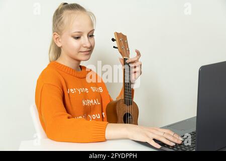 La ragazza adolescente impara a giocare il ukulele e guarda dentro il suo laptop Foto Stock