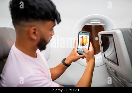 Giovane latino-americano che effettua una videochiamata sul suo cellulare, seduto sull'aereo vicino alla finestra. Foto Stock