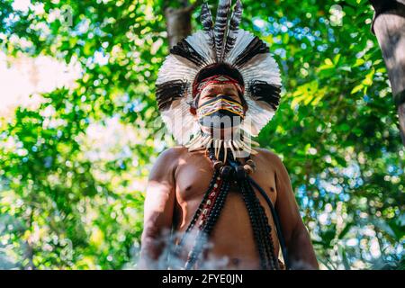 Sciamano della tribù Pataxó. Anziano indiano che indossa headdress di piuma e maschera del viso a causa della pandemia di covid-19. Brasiliano indiano guardando il c Foto Stock