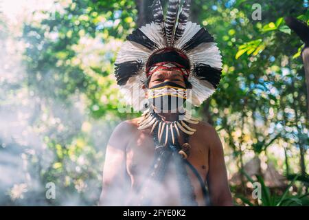 Sciamano della tribù Pataxó. Anziano indiano che indossa headdress di piuma e maschera del viso a causa della pandemia di covid-19. Brasiliano indiano guardando il c Foto Stock