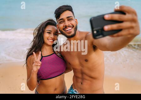 Felice coppia di viaggio facendo selfie sfondo spiaggia, colori estivi soleggiati, umore romantico. Facce emozionali sorridenti felici. Foto Stock