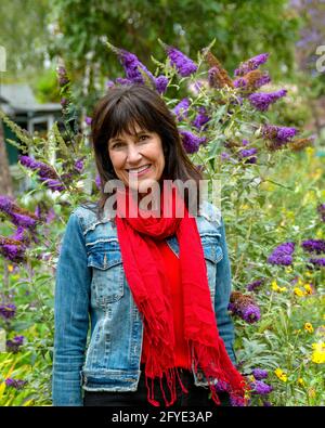 Bella donna anziana moderna e sorridente con spessi capelli brunette, vestita di sciarpa rossa, giacca in denim, si erge di fronte a lussureggianti piante verdi fiorite. Foto Stock