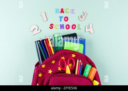 Zaino rosso con diverse coloratissime cancelleria su sfondo verde pastello. Concetto di ritorno a scuola. Vista dall'alto, spazio di copia Foto Stock