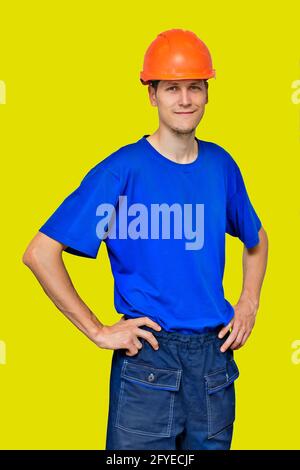 Ritratto di un giovane bell'uomo in un casco da costruzione e abiti da lavoro isolati su uno sfondo giallo. Un lavoratore manuale positivo. Foto Stock