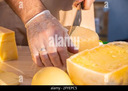 Le mani del negoziante tagliano da vicino una testa di formaggio. Formaggio artigianale in un mercato di strada. Caseificio tagliando il formaggio nel suo laboratorio. Festival del cibo in Foto Stock