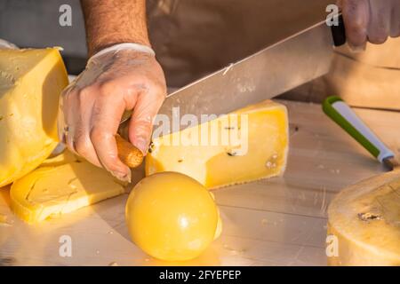 Le mani del negoziante tagliano da vicino una testa di formaggio. Formaggio artigianale in un mercato di strada. Caseificio tagliando il formaggio nel suo laboratorio. Festival del cibo in Foto Stock