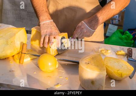 Le mani del negoziante tagliano da vicino una testa di formaggio. Formaggio artigianale in un mercato di strada. Caseificio tagliando il formaggio nel suo laboratorio. Festival del cibo in Foto Stock