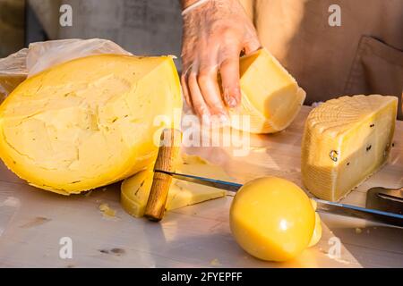 Le mani del negoziante tagliano da vicino una testa di formaggio. Formaggio artigianale in un mercato di strada. Caseificio tagliando il formaggio nel suo laboratorio. Festival del cibo in Foto Stock