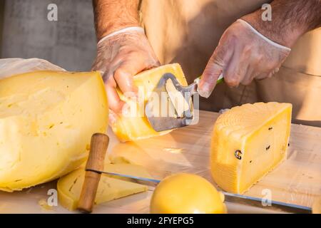 Le mani del negoziante tagliano da vicino una testa di formaggio. Formaggio artigianale in un mercato di strada. Caseificio tagliando il formaggio nel suo laboratorio. Festival del cibo in Foto Stock