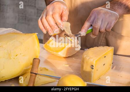 Le mani del negoziante tagliano da vicino una testa di formaggio. Formaggio artigianale in un mercato di strada. Caseificio tagliando il formaggio nel suo laboratorio. Festival del cibo in Foto Stock