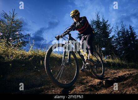 Ampio angolo di vista del giovane uomo che corre in bicicletta in discesa con cielo blu sera sullo sfondo. Cyclist in costume da ciclismo giù per la collina erbosa di notte. Concetto di sport, ciclismo e tempo libero attivo. Foto Stock