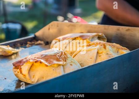 Cibo messicano, un hamburger di bistecca avvolto in una tortilla sotto formaggio fuso sulla griglia di un ristorante all'aperto. Festa del cibo nel parco cittadino. Via f Foto Stock