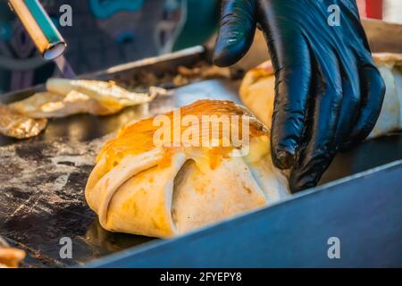 Cibo messicano, un hamburger di bistecca avvolto in una tortilla sotto formaggio fuso sulla griglia di un ristorante all'aperto. Festa del cibo nel parco cittadino. Via f Foto Stock