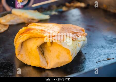 Cibo messicano, un hamburger di bistecca avvolto in una tortilla sotto formaggio fuso sulla griglia di un ristorante all'aperto. Festa del cibo nel parco cittadino. Via f Foto Stock