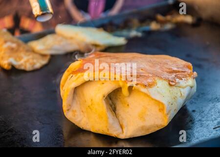 Cibo messicano, un hamburger di bistecca avvolto in una tortilla sotto formaggio fuso sulla griglia di un ristorante all'aperto. Festa del cibo nel parco cittadino. Via f Foto Stock