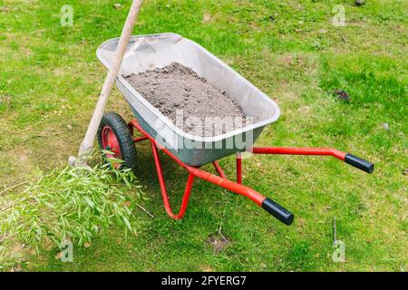 Carrello da giardino con terreno sul prato. Carrello in metallo per lavori in giardino. Utensile da giardiniere con ruote per il trasporto di terra, erbacce, sa Foto Stock