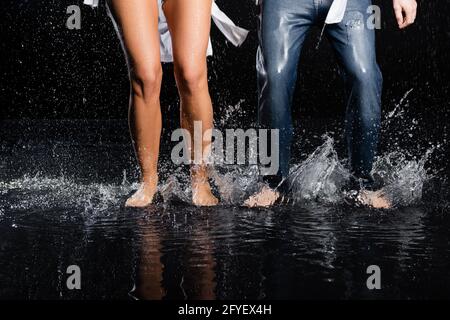 vista parziale delle gambe a piedi nudi maschio e femmina in acqua schizzi su sfondo nero Foto Stock