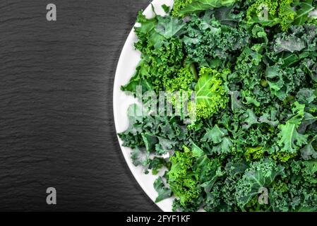 Preparazione di patatine di kale da foglie verdi tagliate su una piastra bianca, vista dall'alto su una lavagna scura Foto Stock