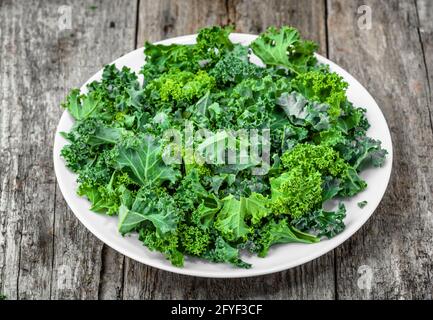 Preparazione di patatine di kale da foglie verdi tritate su piatto bianco, vista dall'alto sfondo rustico Foto Stock