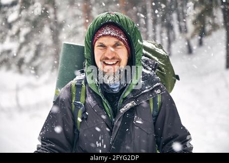 Ritratto di felice arrampicatore con barba e grande zaino in caso di forte nevicata nella foresta di montagna. Concetto di attività all'aperto Foto Stock