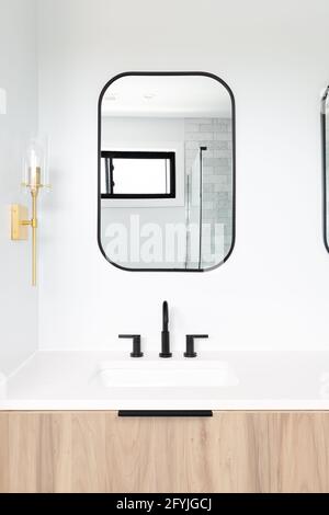 Un bagno splendidamente rinnovato con un armadietto in legno, specchio con cornice nera e rubinetti, e luci dorate montate sulla parete. Foto Stock