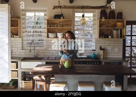 Donna caucasica felice in piedi in cucina cottage utilizzando smartphone e. sorridente Foto Stock