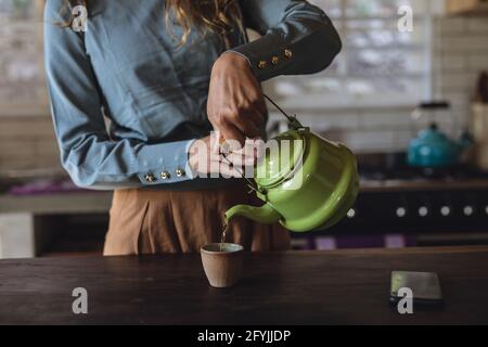 Sezione centrale di donna caucasica in piedi in cucina cottage che versa il tè dalla teiera Foto Stock