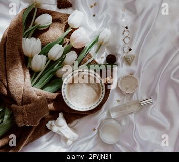 Mazzo di tulipani bianchi avvolti in un maglione di lana su una tovaglia di seta con candele, profumo e gioielli Foto Stock