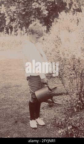 Cartolina fotografica d'epoca 1911 che mostra un ragazzo che annaffiava i fiori in un giardino con la sua lattina d'annaffiatura. Foto Stock