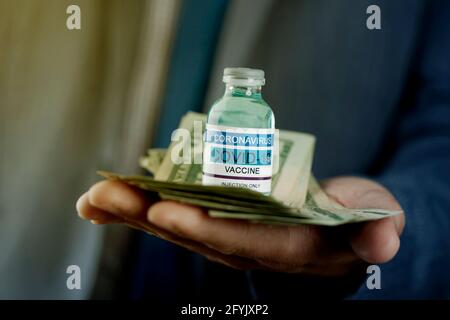 un uomo d'affari, che indossa una giacca blu-grigiastro e una cravatta blu, ha una bottiglia simulata di vaccino covid-19 e un mazzo di banconote in dollaro nelle sue mani Foto Stock