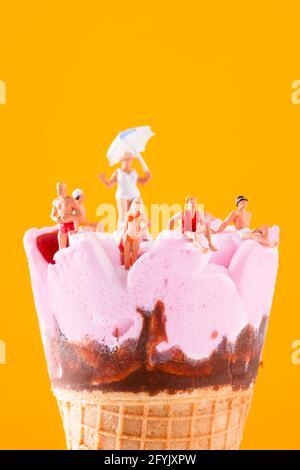 un gruppo di persone in miniatura che indossano un costume da bagno rilassante su un cono rosa pre-riempito di gelato, su uno sfondo arancione con un po' di spazio vuoto sulla parte superiore Foto Stock