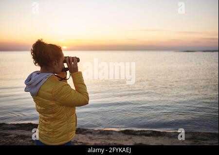 Vista laterale di una giovane donna con giacca gialla guardando attraverso il binocolo e godendo di una splendida vista del paesaggio costiero Foto Stock