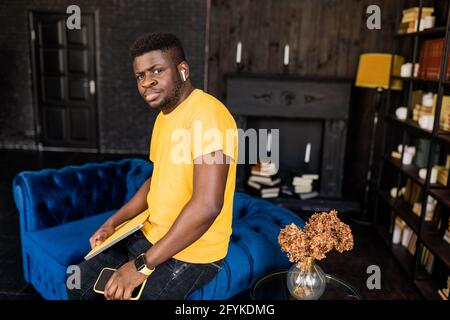 Imprenditore africano-americano serio millennial di affari uomo in piedi a casa a un divano elegante, guardando con sicurezza la fotocamera, tenere giallo notebook e. Foto Stock