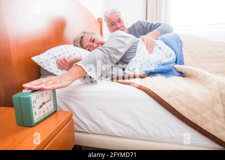 Felice coppia senior a casa ferma la sveglia al risveglio mattutino - la donna tocca il colck per continuare a dormire l'uomo che la abbracciava - luce finestrino interna persone anziane l Foto Stock