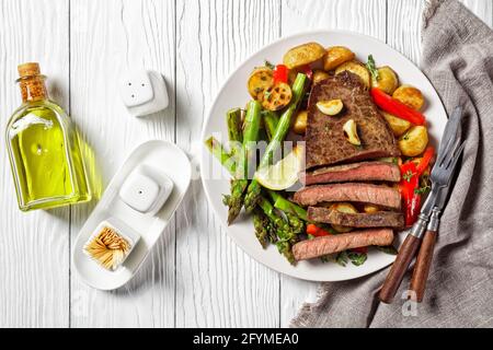 bistecca di manzo con patate novelle arrostite, asparagi, limone e pepe rosso su un piatto bianco con salsa al barbecue su un tavolo di legno, piatto Foto Stock