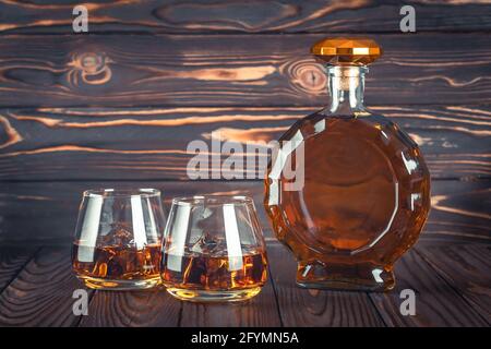 Una bottiglia di whisky e due bicchieri su un tavolo di legno marrone scuro. Bevanda alcolica gialla trasparente con ghiaccio. Brandy, bourbon. Alcool forte bevuto Foto Stock