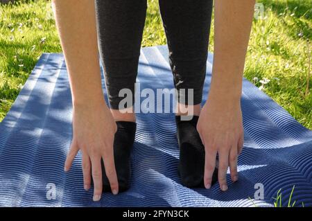 Donna che indossa leggings grigi e calze nere, pratica yoga e stretching su un tappeto blu sull'erba verde all'aperto Foto Stock