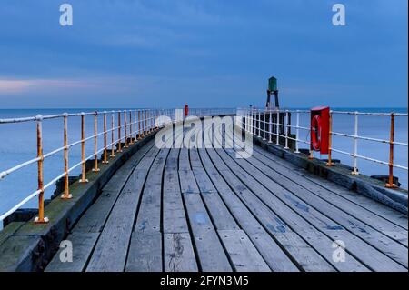 Whitby's West Pier (mare calmo, passerella in legno, faro, spettacolare luce notturna blu dell'ora) - ingresso al porto di Whitby, costa del Nord Yorkshire, Inghilterra Regno Unito Foto Stock