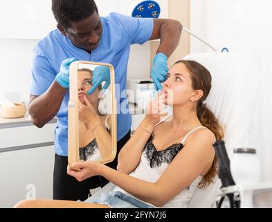 Cosmetologa maschile che dà consultazione alla paziente giovane donna sdraiata sul divano in clinica estetica Foto Stock