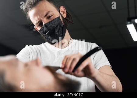 Il rasoio dritto taglia la barba mans in barbershop. Barbiere uomo in maschera medica facendo acconciatura per l'uomo bello Foto Stock