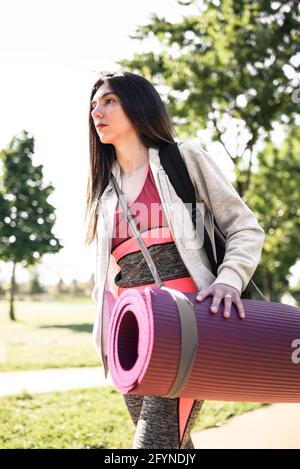 Donna caucasica su abiti sportivi a piedi su un parco verde. Porta con sé un tappeto di yoga viola Foto Stock