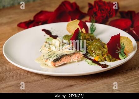 Delizioso filetto di pesce rosso con salsa bianca con verdure in salsa verde su un piatto bianco. Un bel piatto da ristorante decorato con petali di rosa. Foto Stock