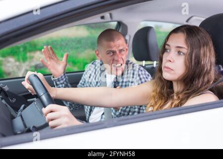 Giovane donna confusa che guida l'auto sullo sfondo di un uomo arrabbiato che urla seduto sul sedile del passeggero Foto Stock